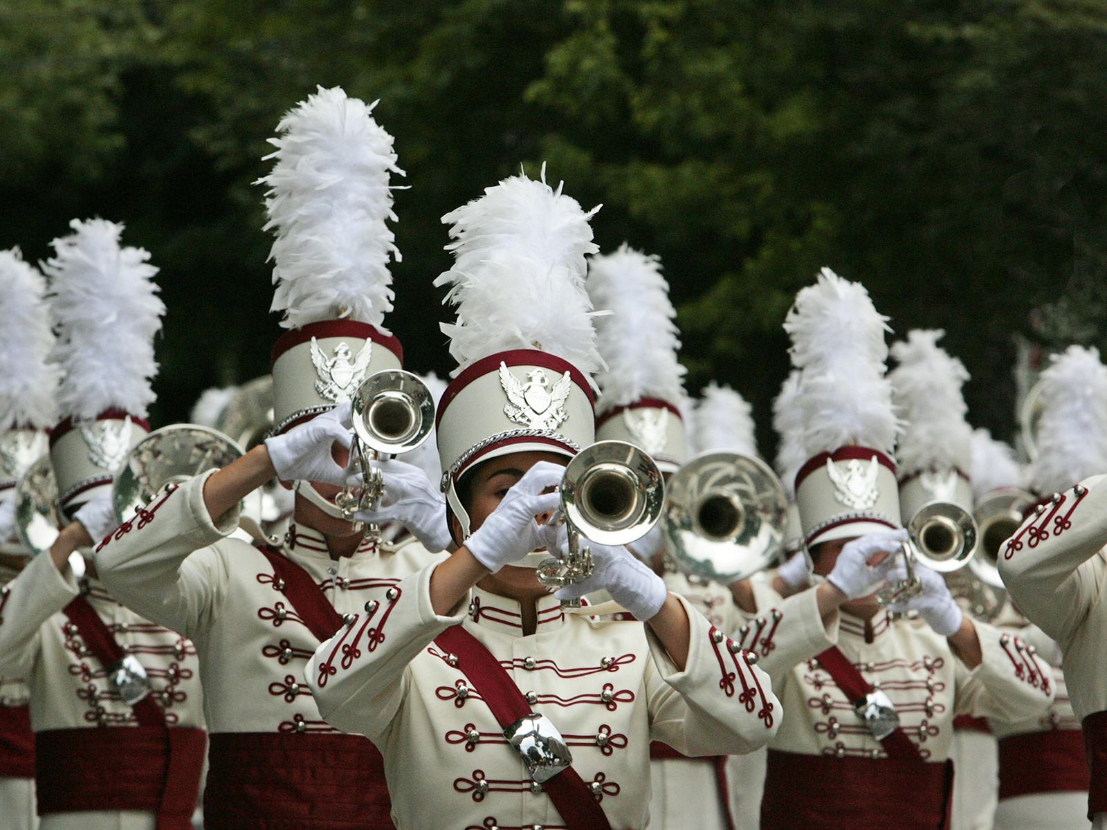 Marching band members in white and maroon uniforms with plumed hats playing trumpets